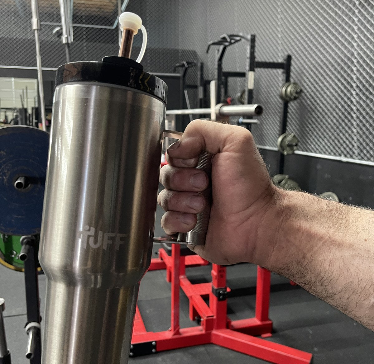 A hand wrapped around the knurled barbell handle of the 40 oz Tuff Tumbler at a squat rack.
